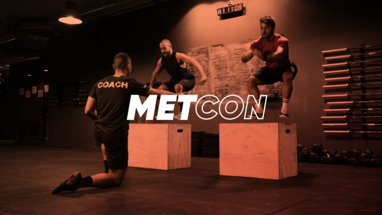 Man wearing a shirt with the word "Coach" on his back, training 2 clients as they workout doing box jumps