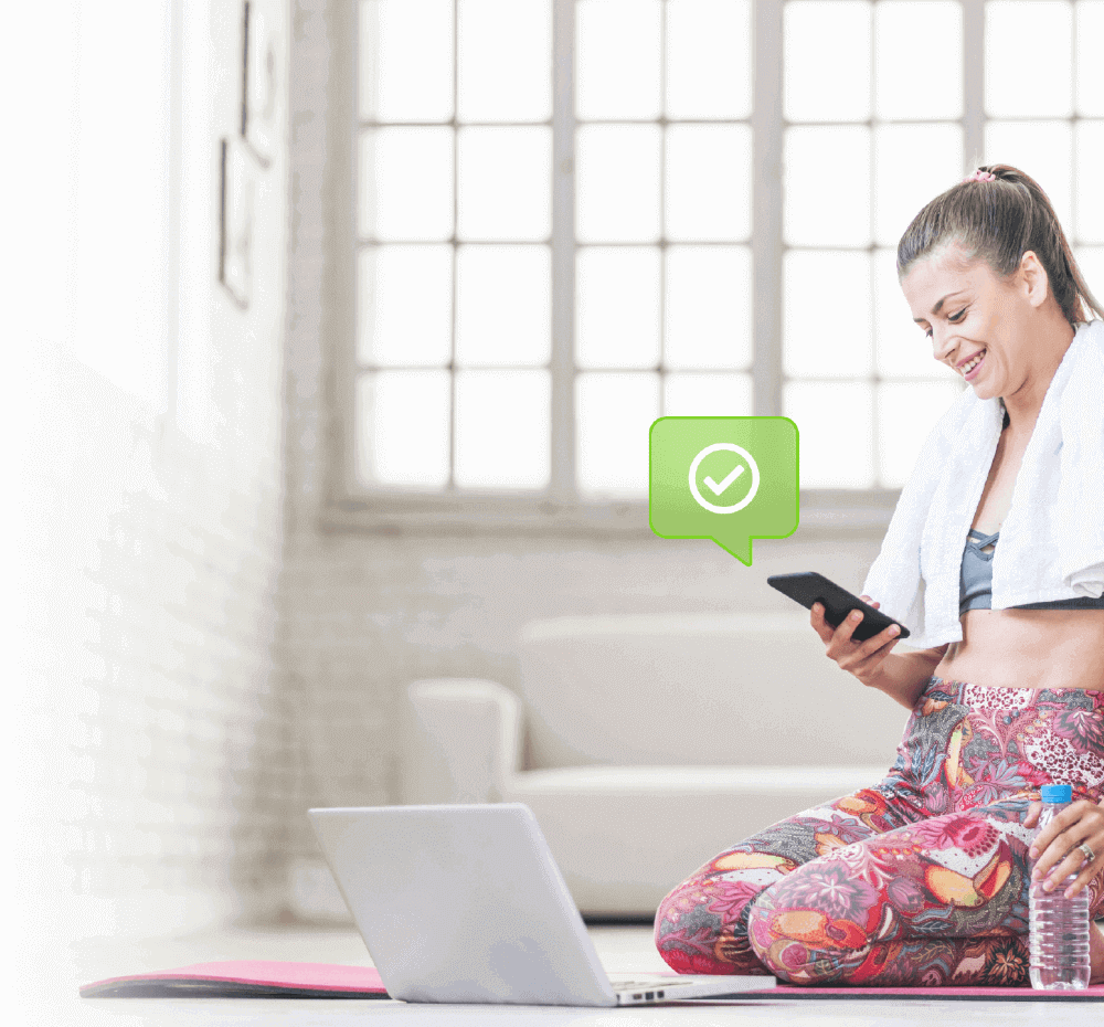 Fit woman sitting in front of laptop holding a water bottle and looking at her phone smiling