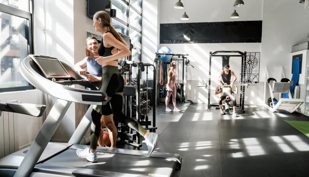 Cheerful female athlete in sportswear exercising on treadmill while talking to male instructor during fitness workout in modern gym