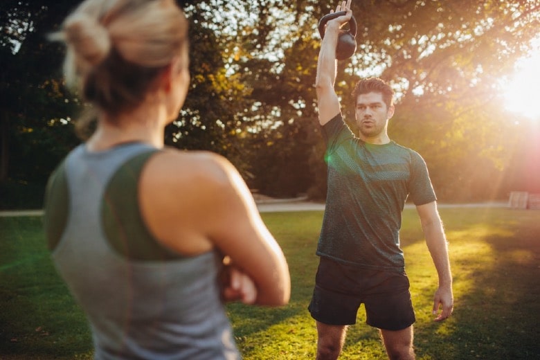 Healthy young man with female trainer exercising with kettlebell in the park. Fit man exercising with female trainer in front.