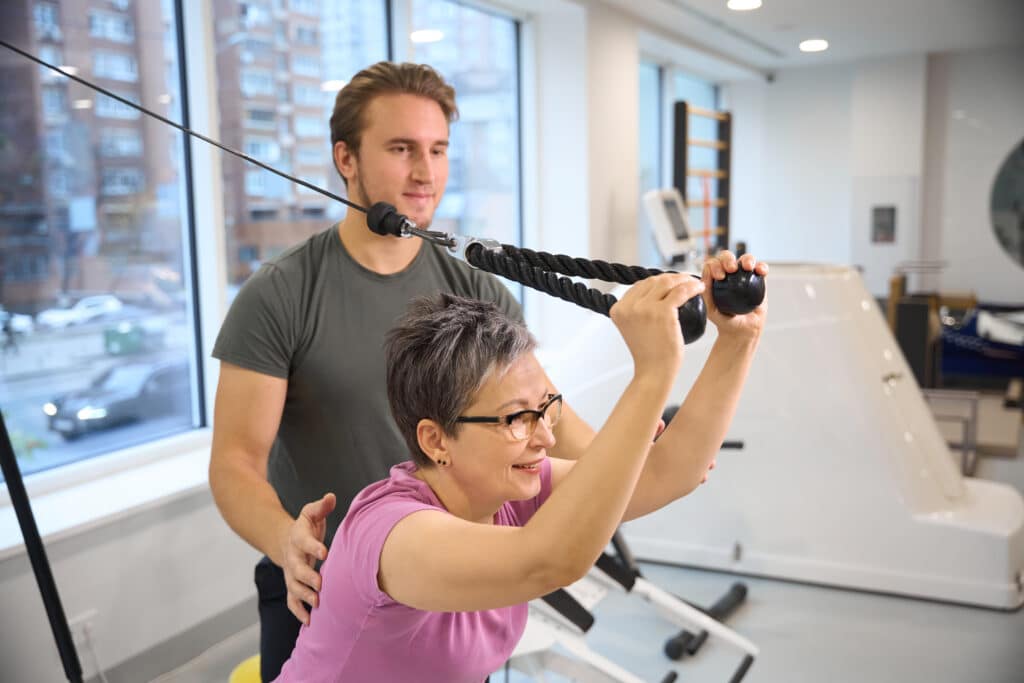 Male trainer teaching smiling adult caucasian woman wearing sportswear doing sport exercise for triceps in gym.
