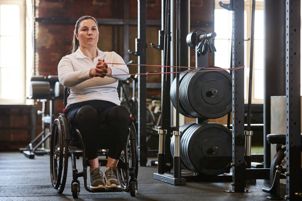 Full-length portrait of adult woman with disability training with elastic bands in gym.