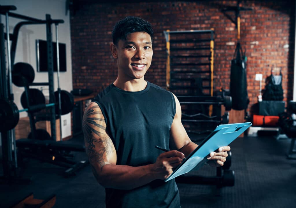 Successful male personal trainer with a tattooed arm holding a clipboard and smiling in a brick-walled gym with weightlifting equipment.
